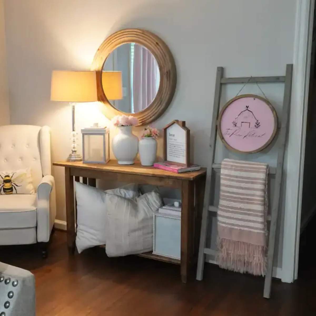 A cozy corner of a room features a white armchair beside a wooden console table, adorned with decorative items and a round mirror, with a ladder displaying a woven blanket.