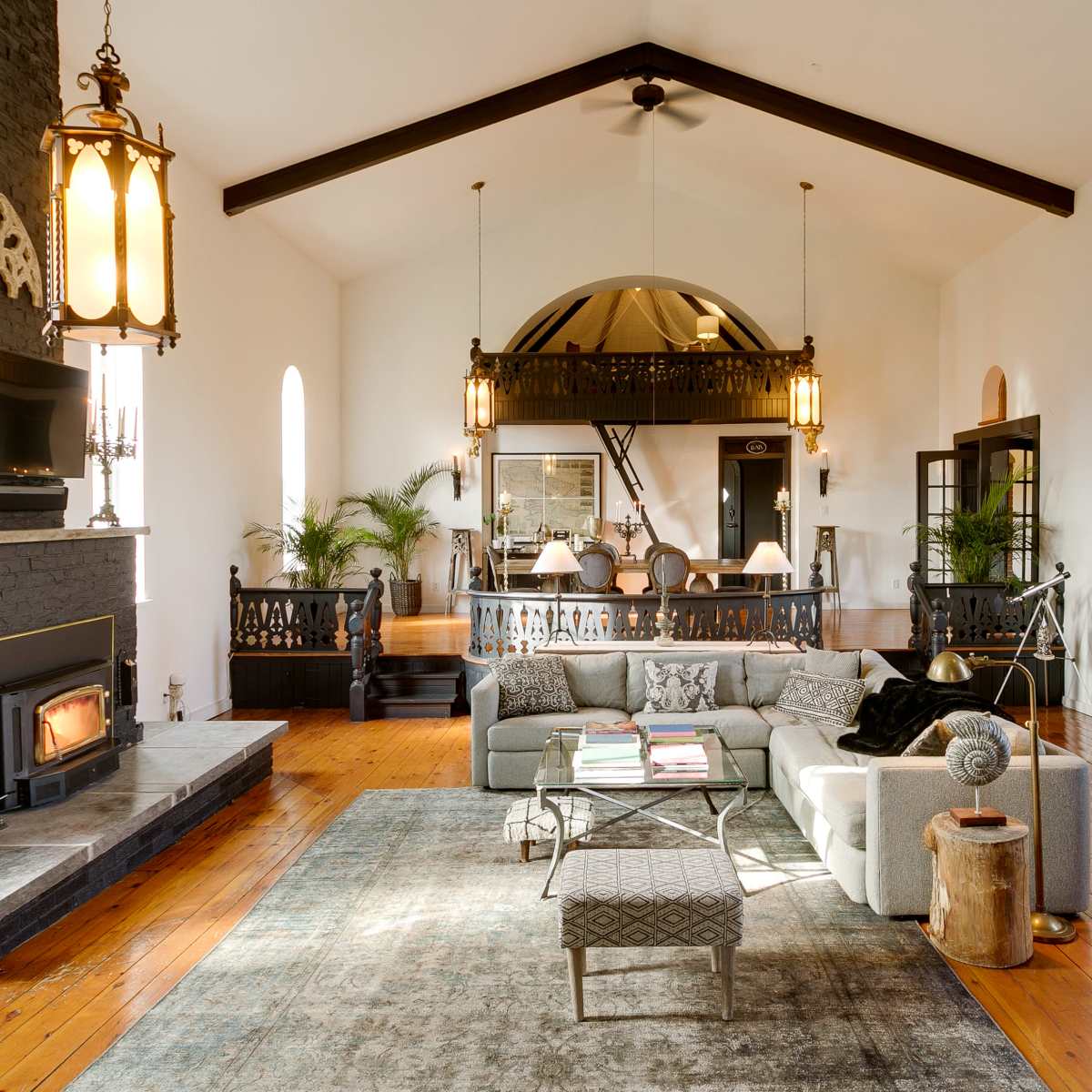 A spacious living room features high ceilings, large windows, a fireplace, and a mix of modern and traditional furnishings.