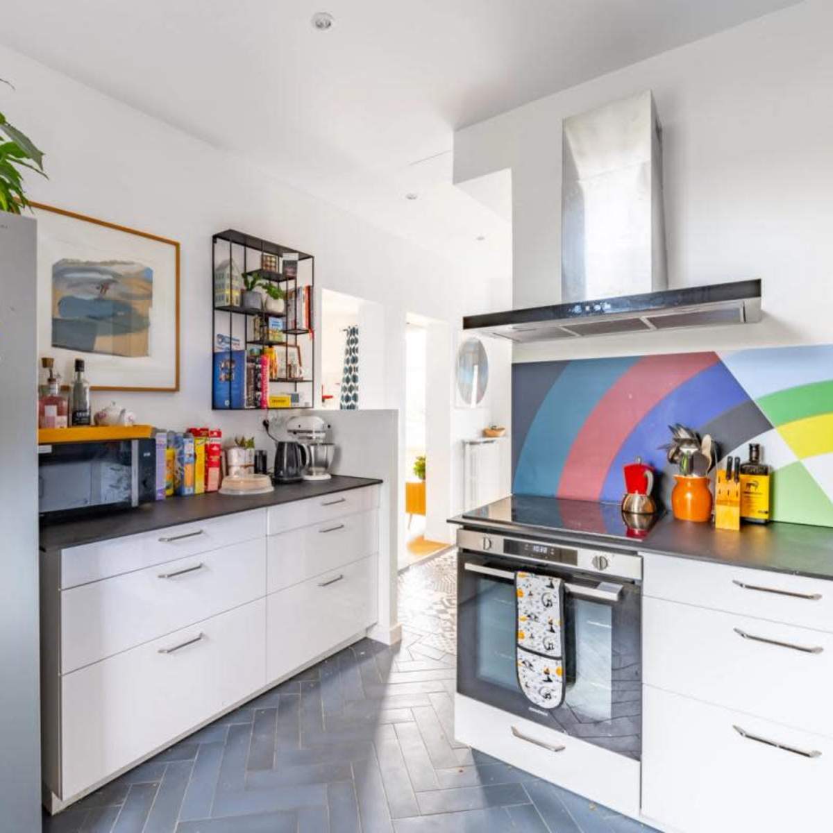 A modern kitchen with white cabinetry, a colorful backsplash, stainless steel appliances, and a vibrant decorative element on the wall.