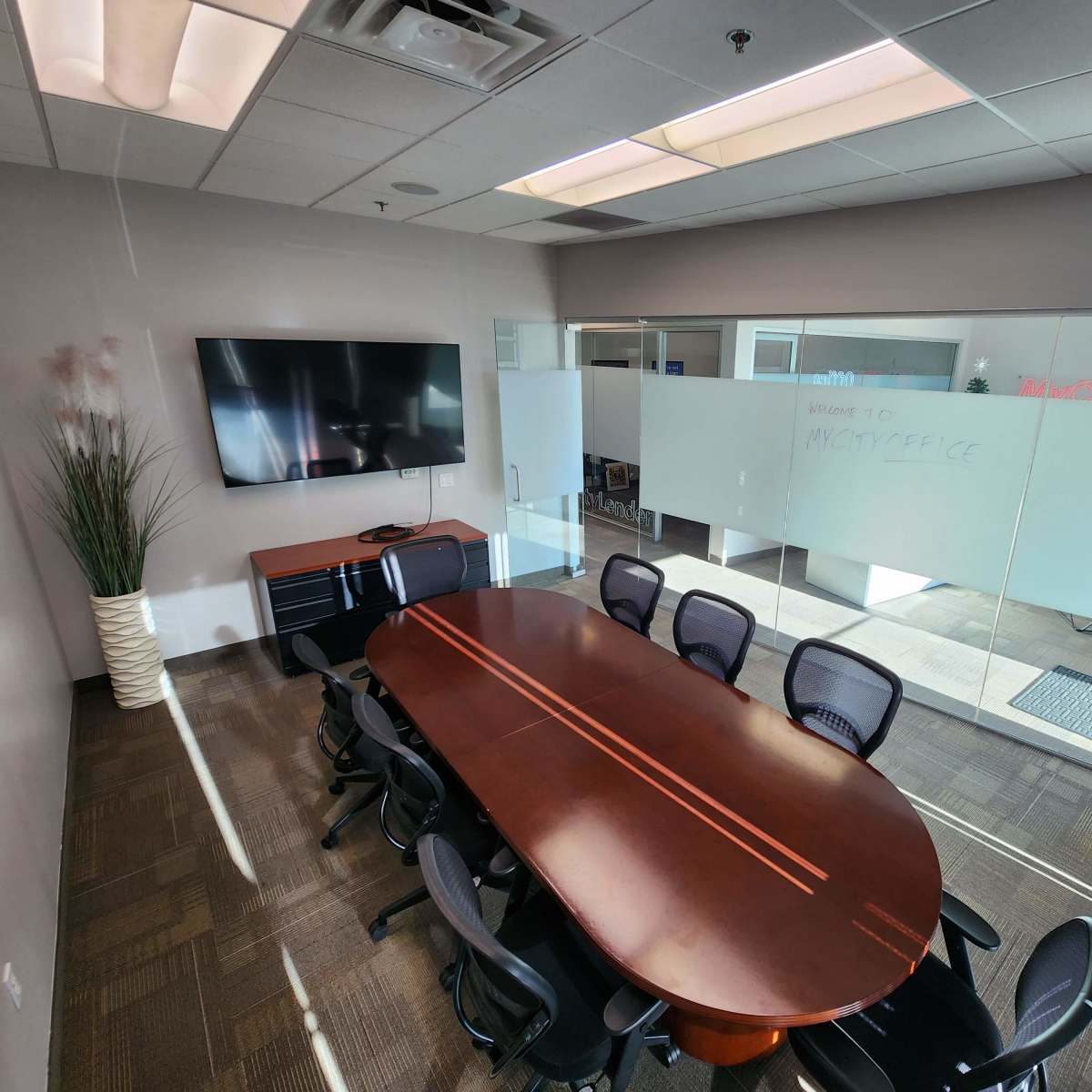 A conference room featuring a large oval wooden table surrounded by black chairs, a wall-mounted TV, and glass walls with whiteboards on one side.