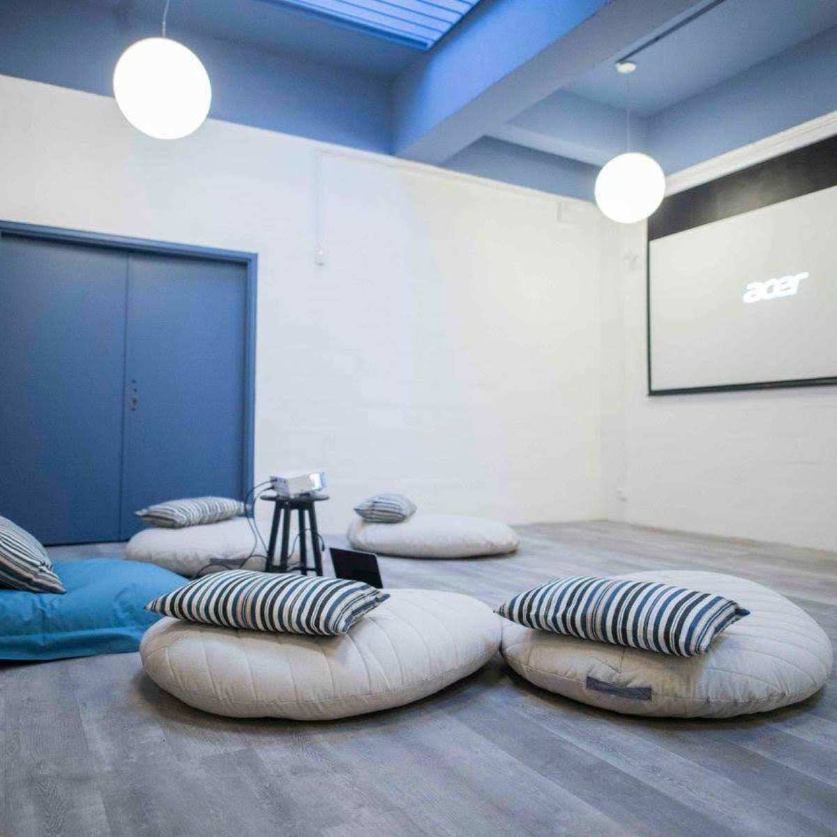A minimalist room features several large floor cushions, a small table, and a projector screen on a white wall.