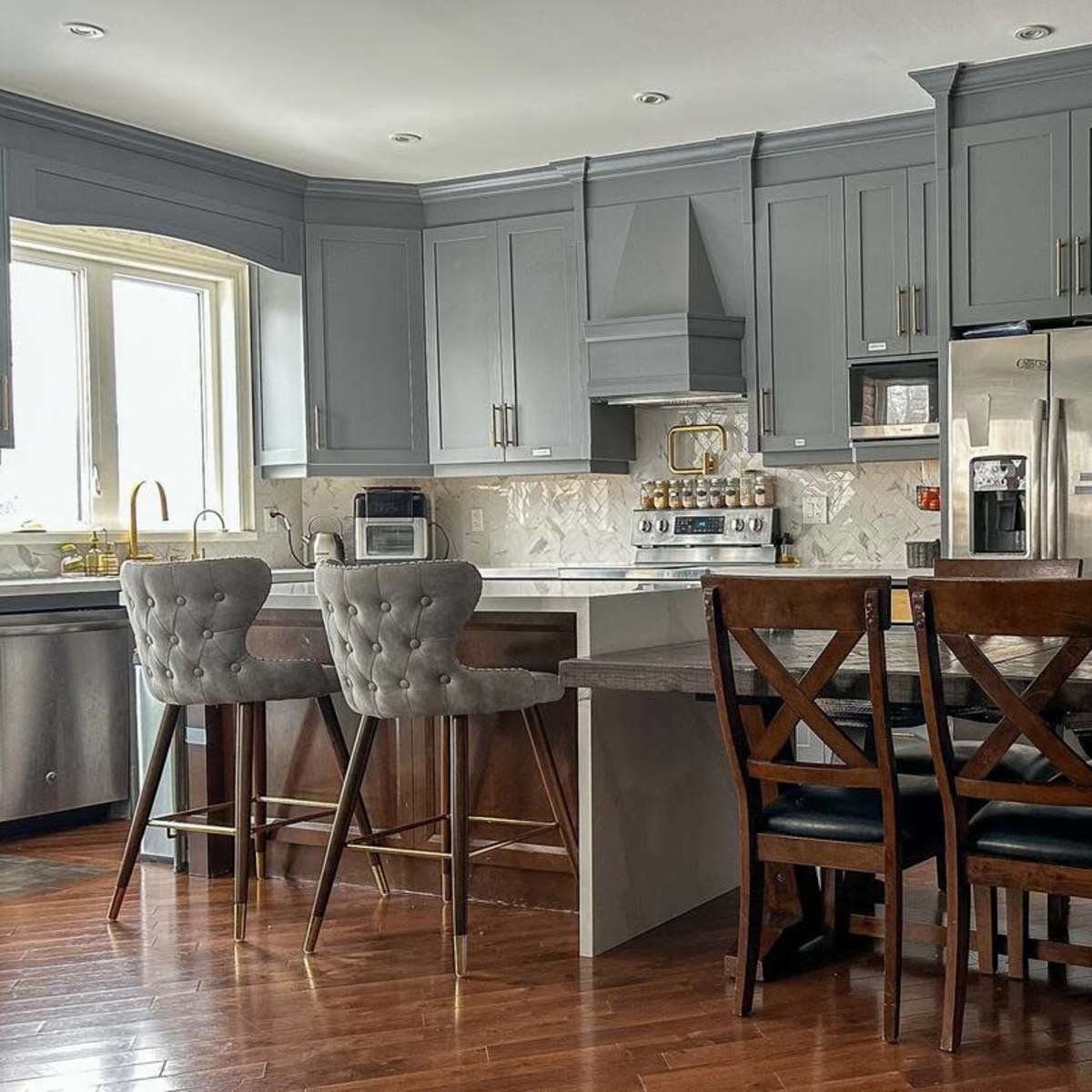 A modern kitchen with gray cabinets, a stainless steel refrigerator, a marble backsplash, and a wooden dining table surrounded by chairs.