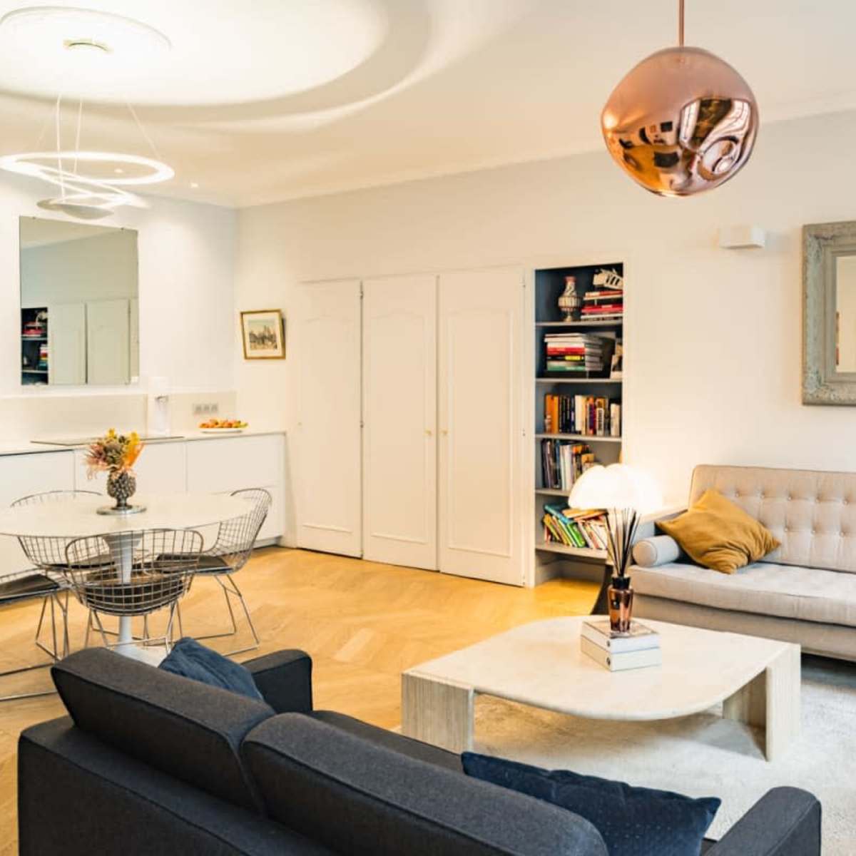 A modern living room features a light-colored sofa, a dark sofa, a round dining table with wire chairs, and a coffee table, all complemented by a mirrored wall and decorative lighting.