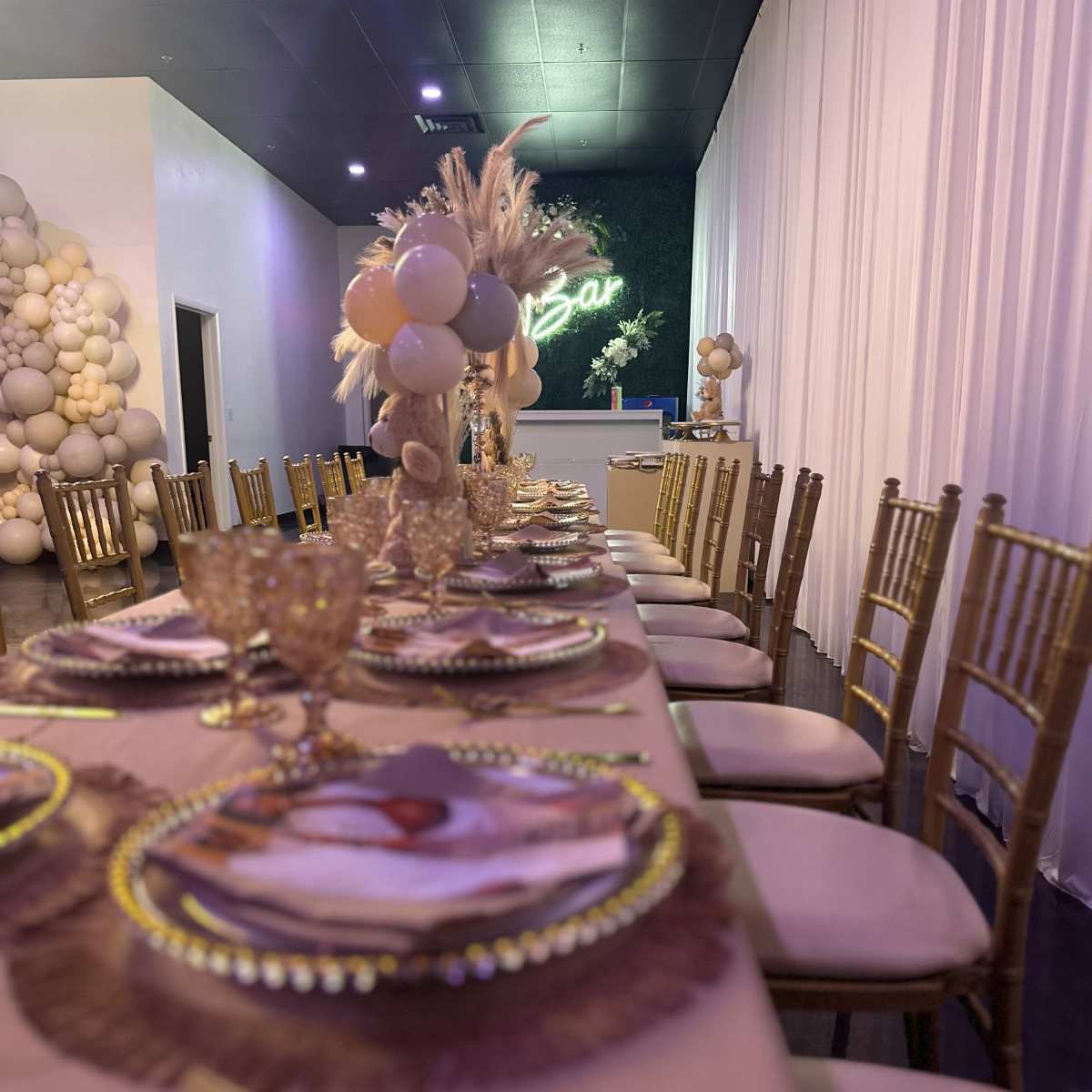 A long dining table set with elegant place settings, surrounded by gold chiavari chairs, and decorated with a floral centerpiece and balloons against a softly lit backdrop.