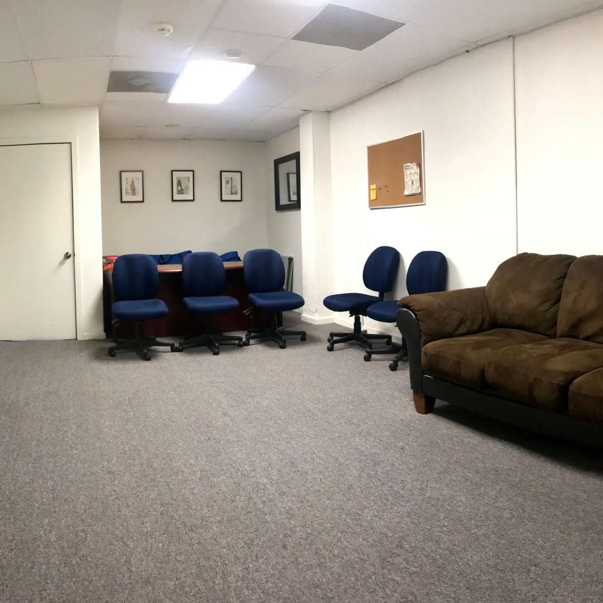 A sparsely furnished room with a brown couch, several blue chairs arranged around a table, a television on the wall, and a bulletin board.