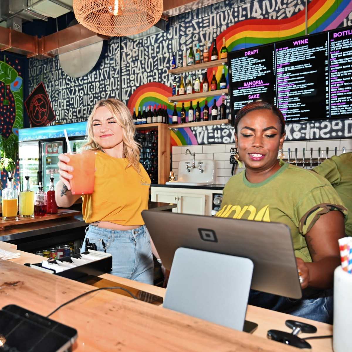 A bar scene features two women, one serving a colorful drink and the other operating a tablet, set against a vibrant mural and a menu board.