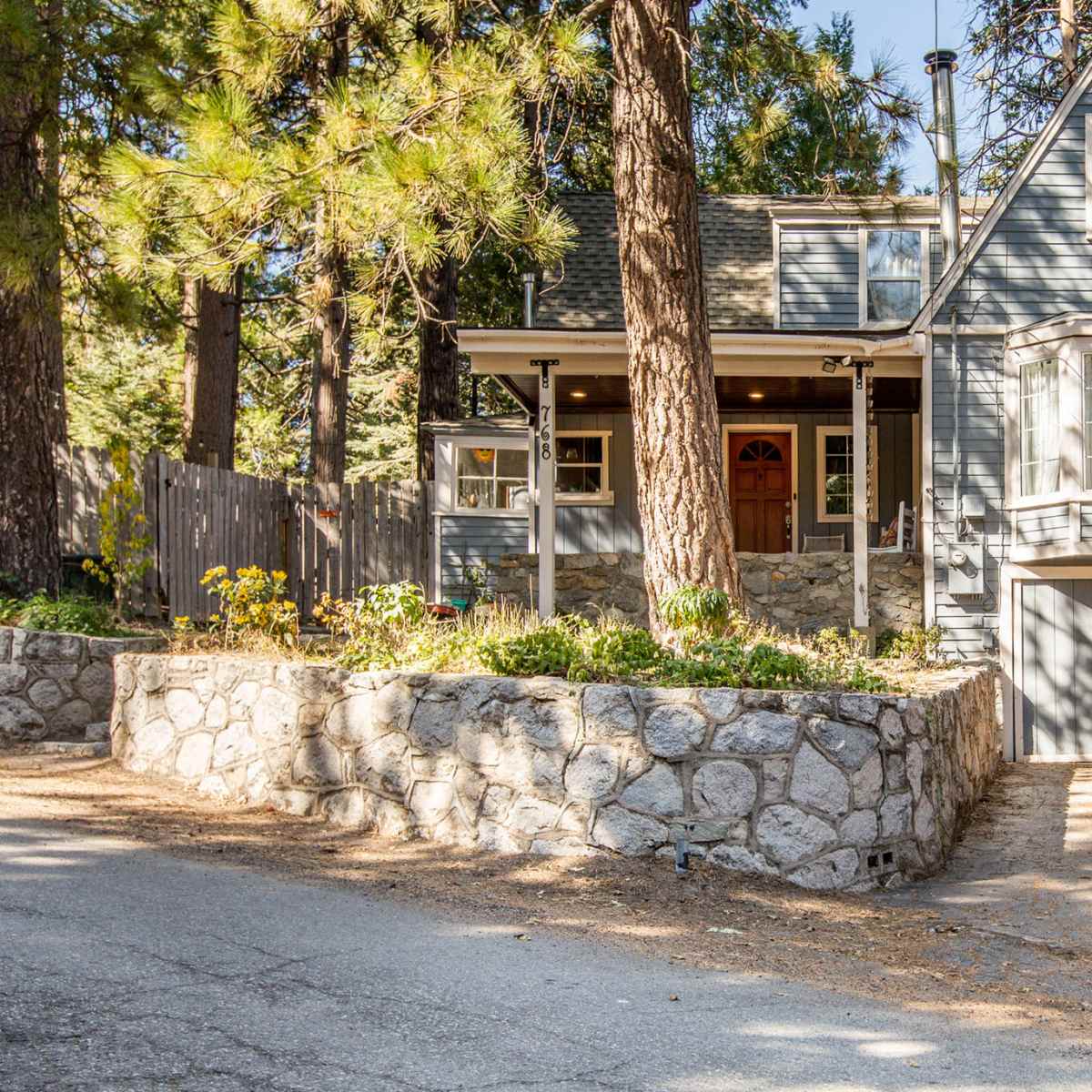 A gray two-story house with a stone foundation is situated on a tree-lined street.