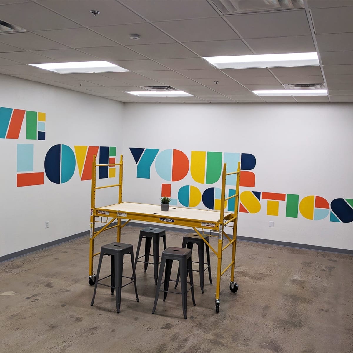 An empty office space with a yellow scaffold and three black stools in front of a colorful wall mural that reads "WE LOVE YOUR LOGISTICS."