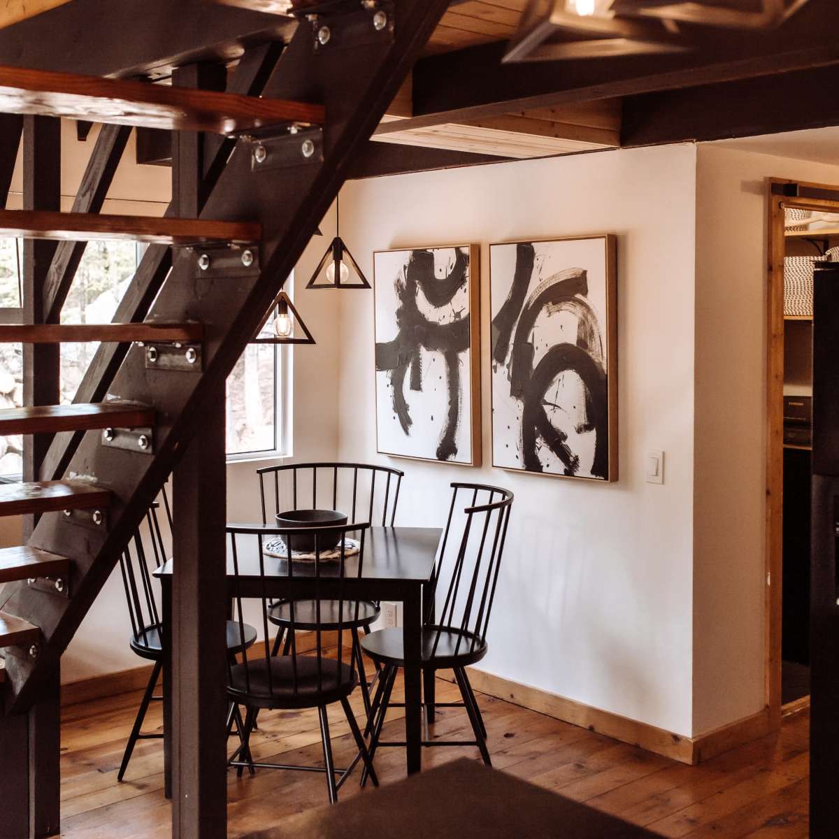 A dining area with a black table and chairs beneath abstract wall art, adjacent to a staircase with wooden steps and metal railing.