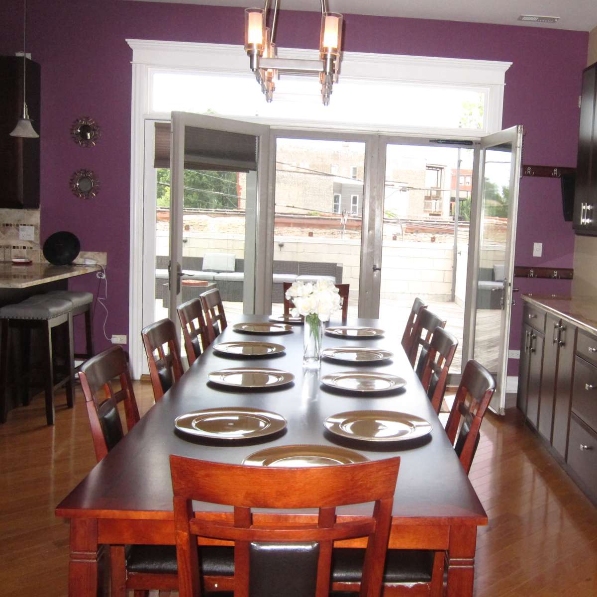A long wooden dining table with plates set is positioned in a brightly lit kitchen, featuring purple walls and large double doors that open to an outdoor area.