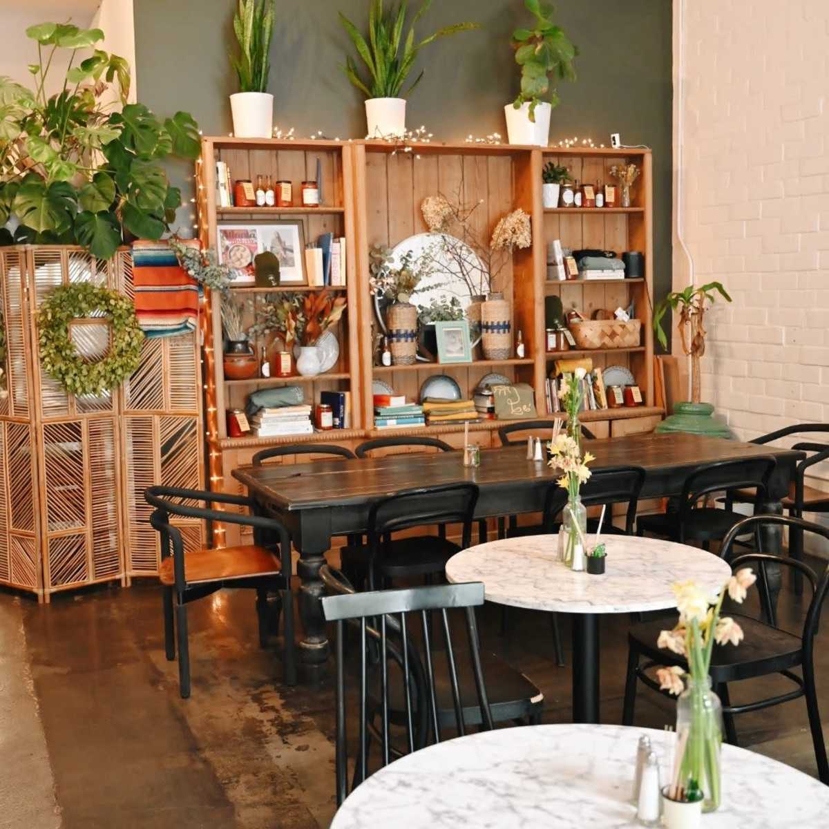 A cozy café interior featuring wooden and marble tables, shelving filled with decorative items, and greenery accenting the space.