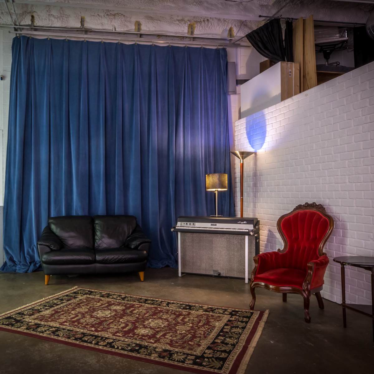 A studio room with a dark blue curtain, a black leather couch, a red armchair, a side table, and a vintage keyboard on a gray surface.