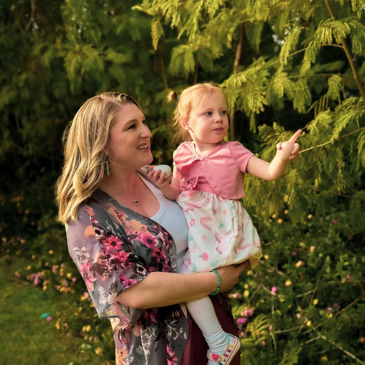 A woman holds a young girl outdoors, surrounded by greenery and flowers.