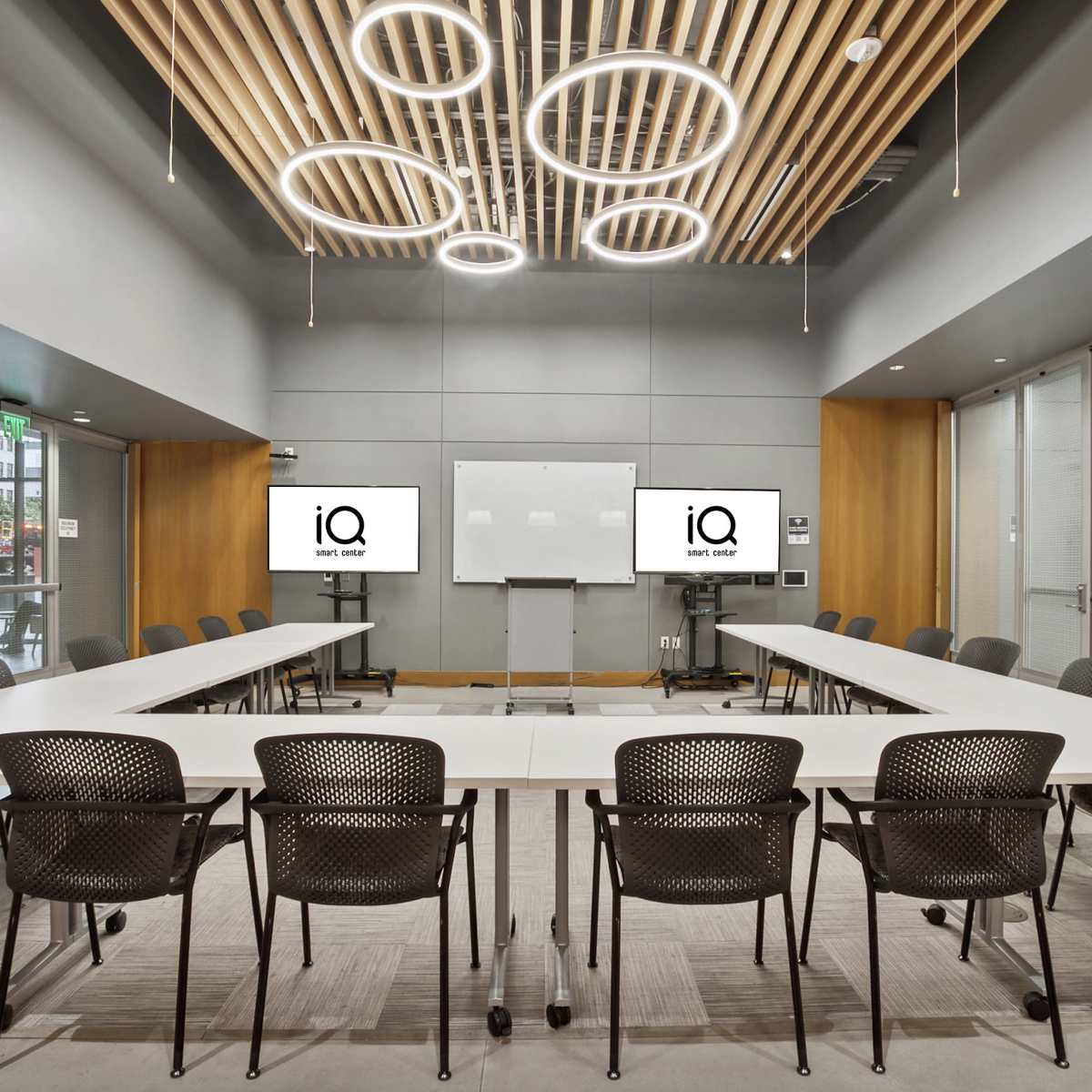 A modern conference room features a large oval table surrounded by black chairs, with two screens displaying the logo "iQ" and a whiteboard on the wall.