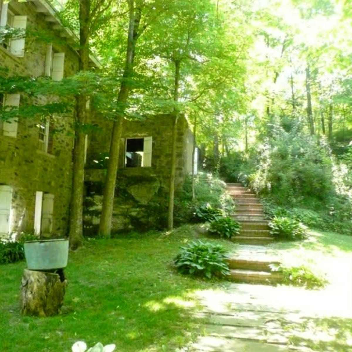 A stone building with white shutters is surrounded by trees and a grassy area, leading to a set of stone steps.