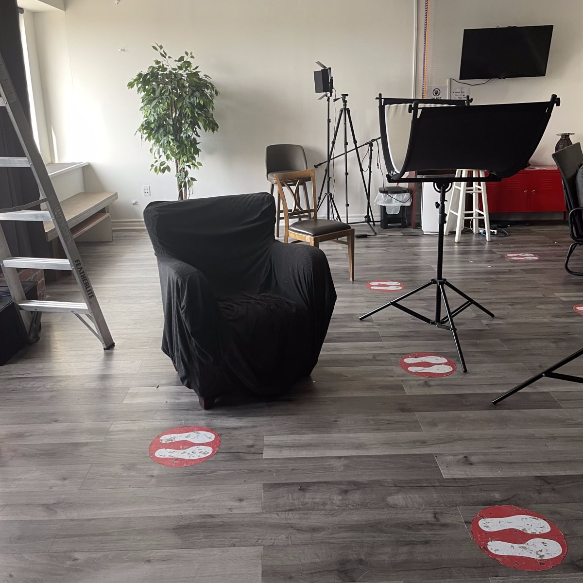 A photo studio with a chair covered in black fabric, a ladder, lighting equipment, a monitor, and marked positions on the floor.