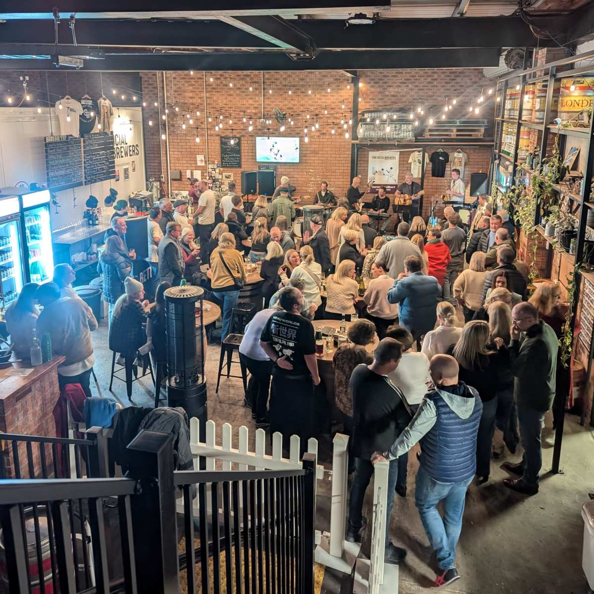 A crowded indoor venue features numerous people socializing and sitting at tables, with a large screen displaying a sports event in the background.