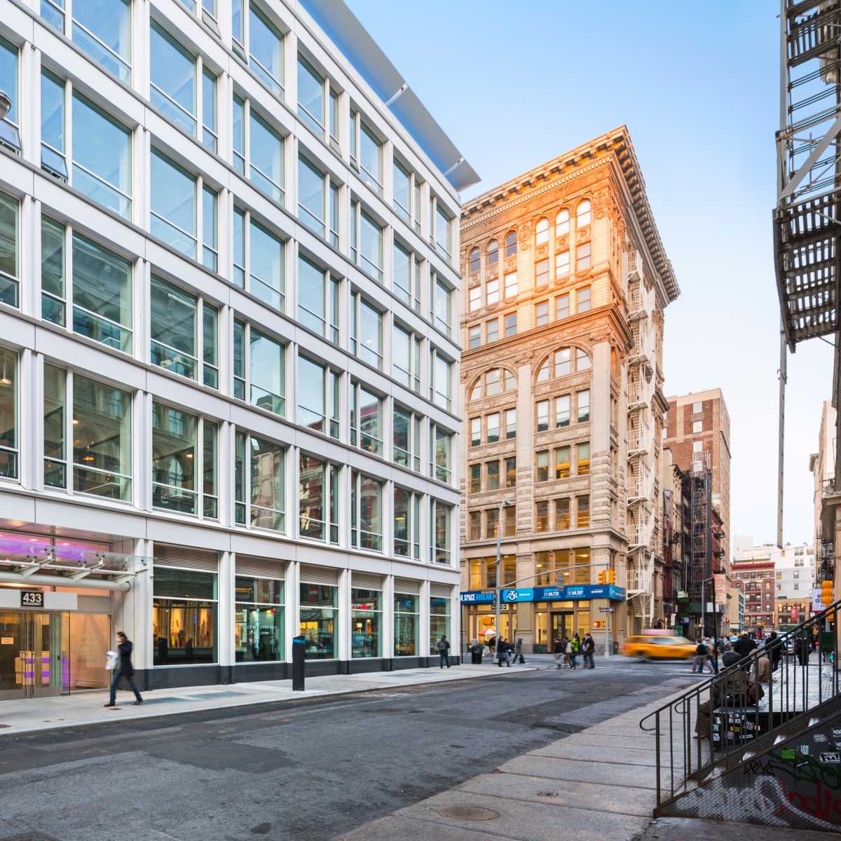 A modern glass-fronted building stands at the intersection of a city street, flanked by older brick structures.