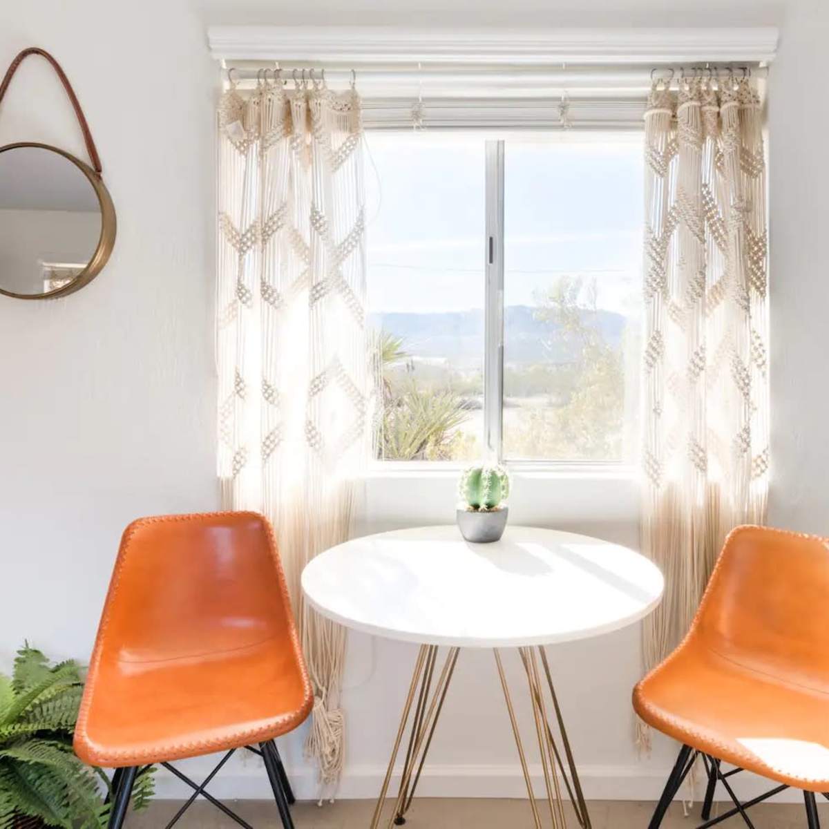 A small dining area features a round table with two orange chairs, a plant in the center, and a window showing a mountainous landscape.