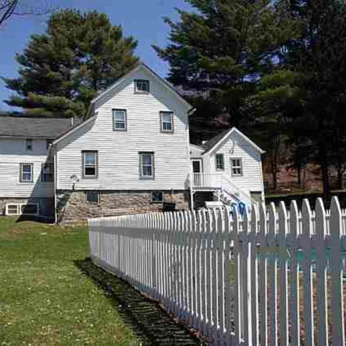 A two-story white house with a stone foundation, surrounded by a white picket fence and green lawn, under a clear blue sky.