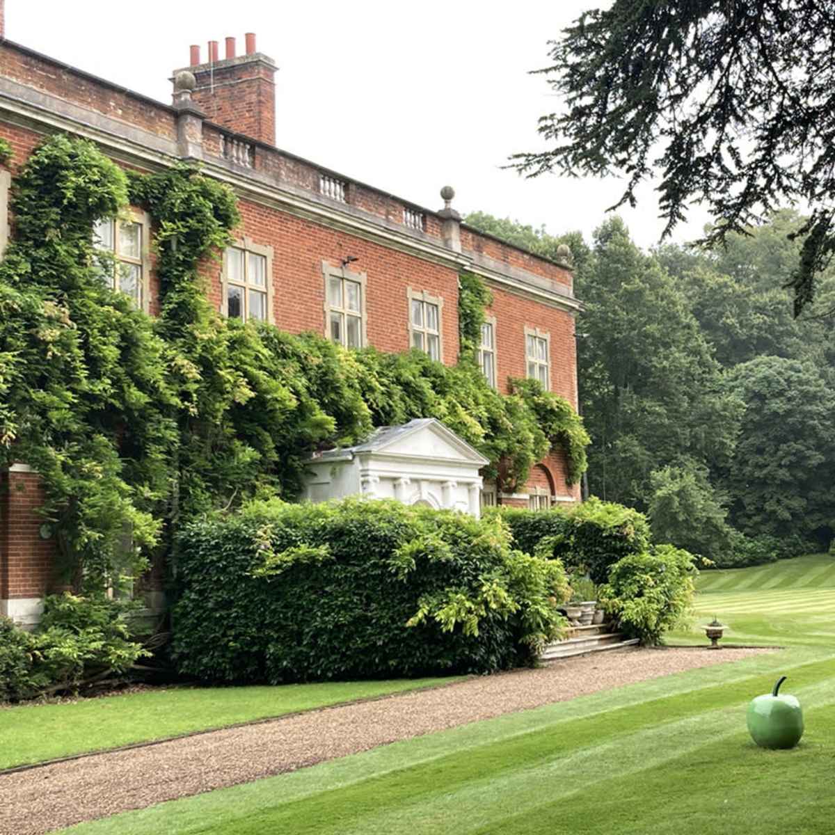 A large brick mansion with ivy covering its facade is set against a neatly manicured lawn featuring a giant green apple sculpture.