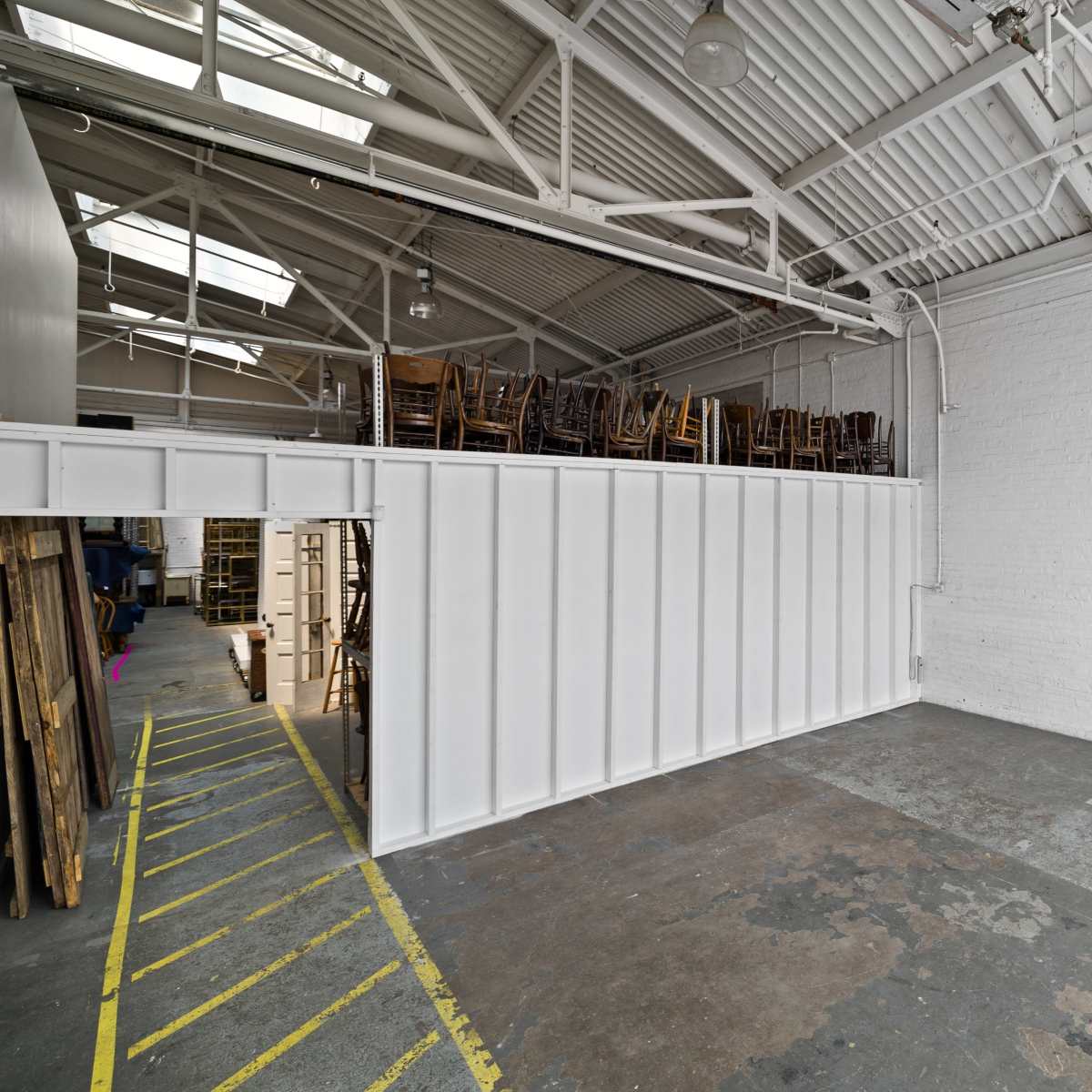 A spacious, empty warehouse interior with a stack of wooden chairs on a raised platform against a white wall.