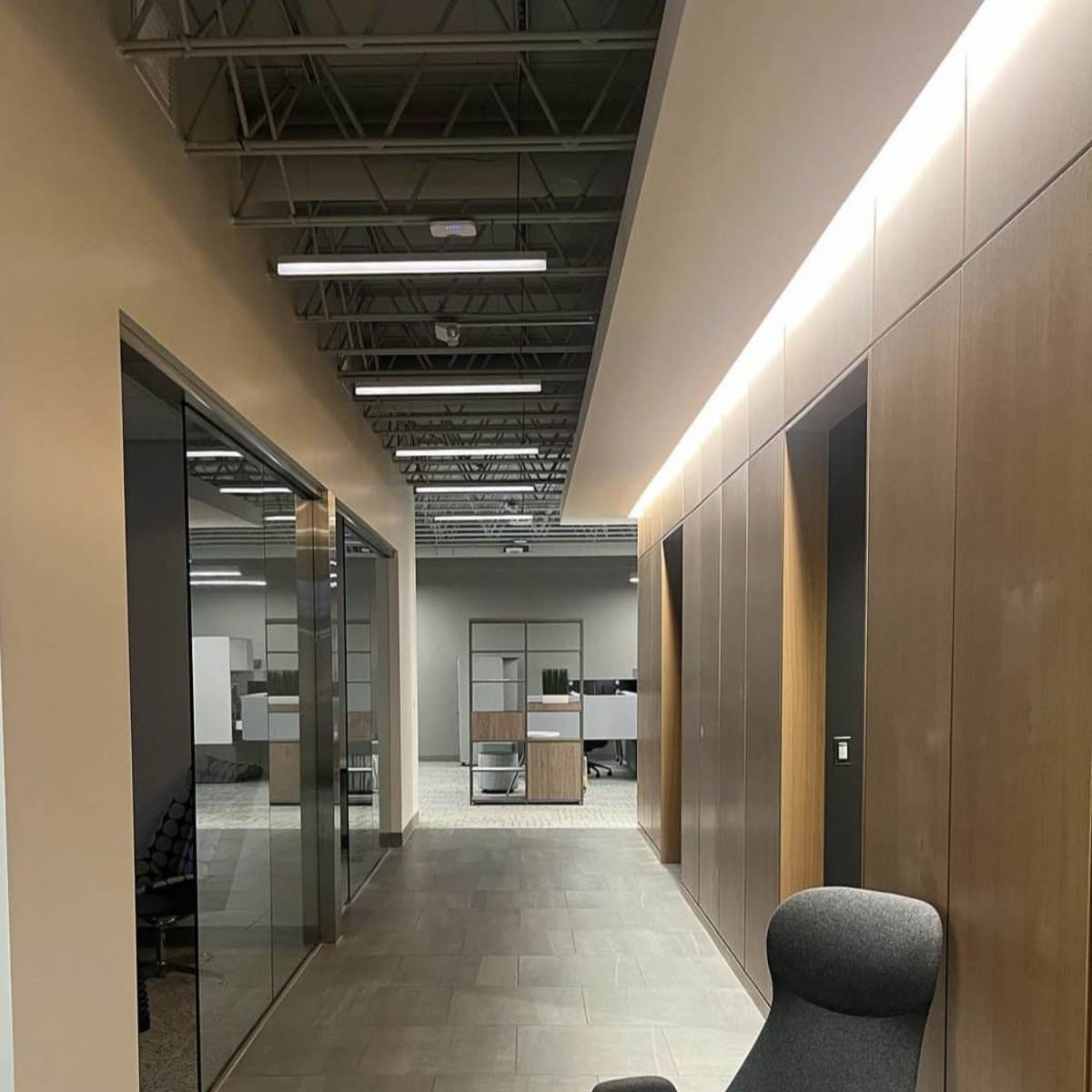 A modern office hallway with a gray chair positioned next to glass partitioned rooms and wooden paneling on the walls.