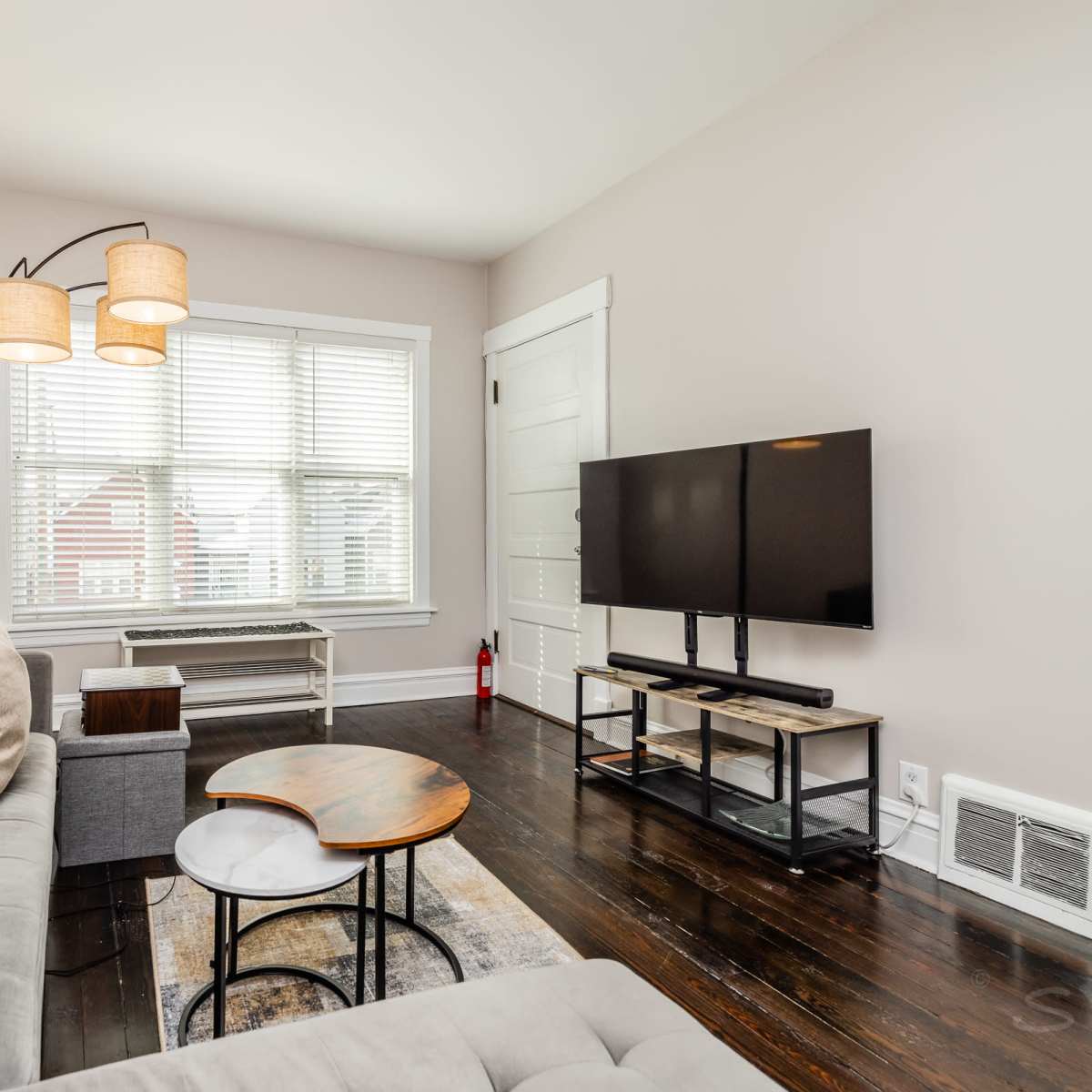 A living room with a gray sofa, a wooden coffee table, a floor lamp with two shades, a television on a stand, and a window with white blinds.