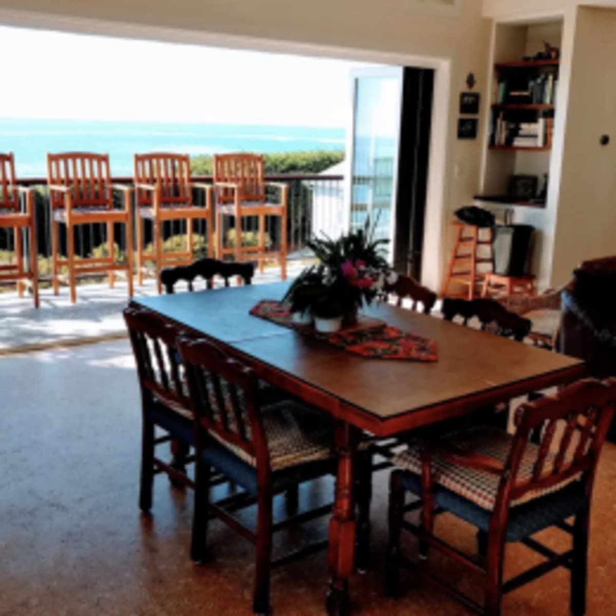 A dining table with eight chairs is positioned indoors overlooking a patio with bar stools and a view of the water.