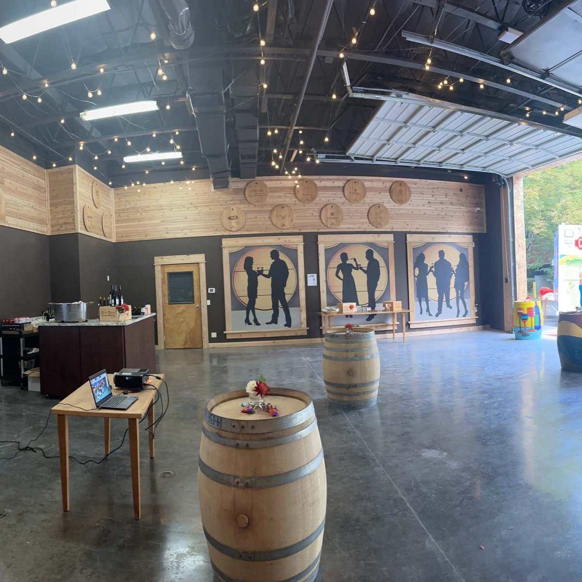 The interior of a rustic event space features wooden barrel tables, a large projection screen, and silhouette artwork on the walls.