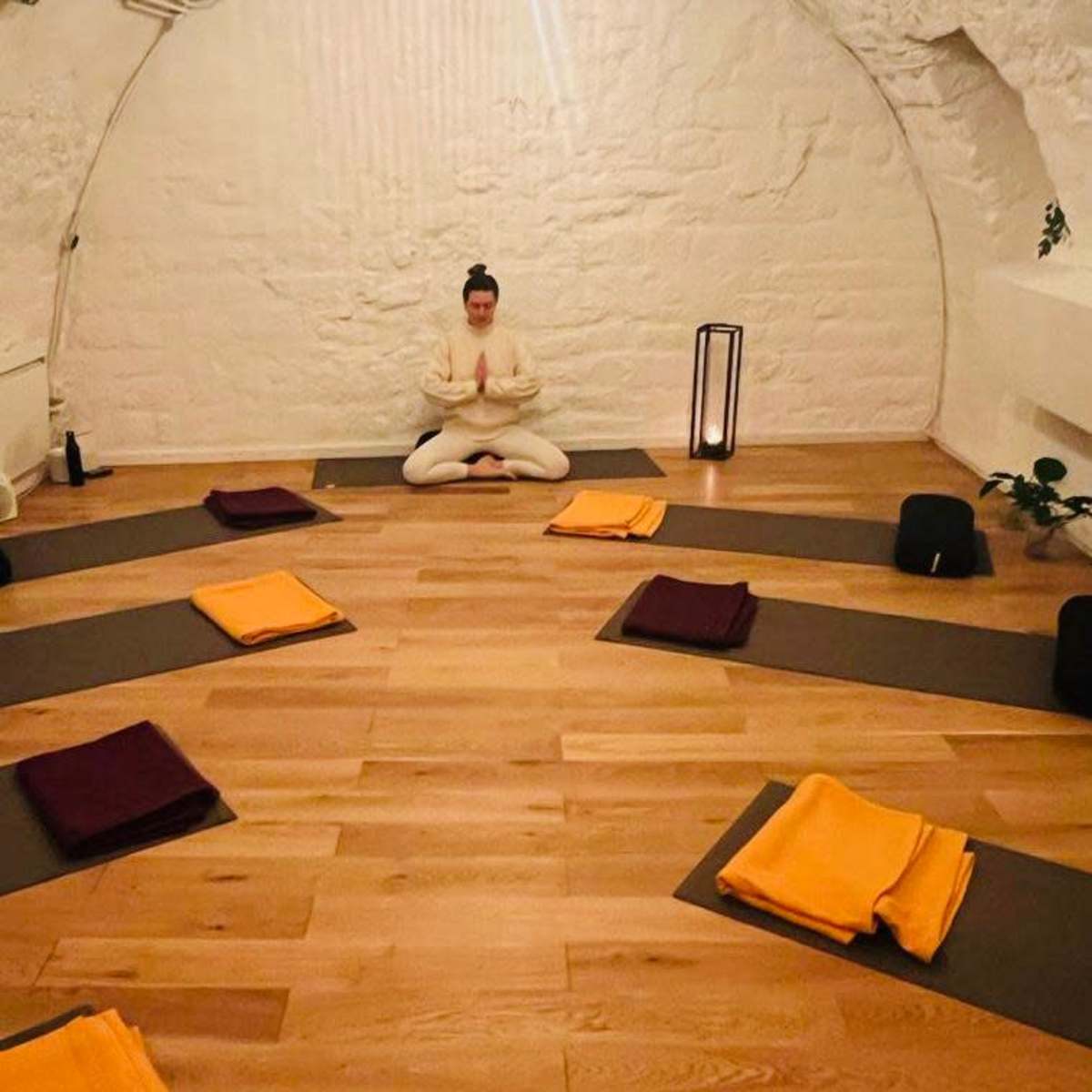 A person sits in a meditative pose surrounded by neatly arranged yoga mats and blankets in a serene, softly lit room.