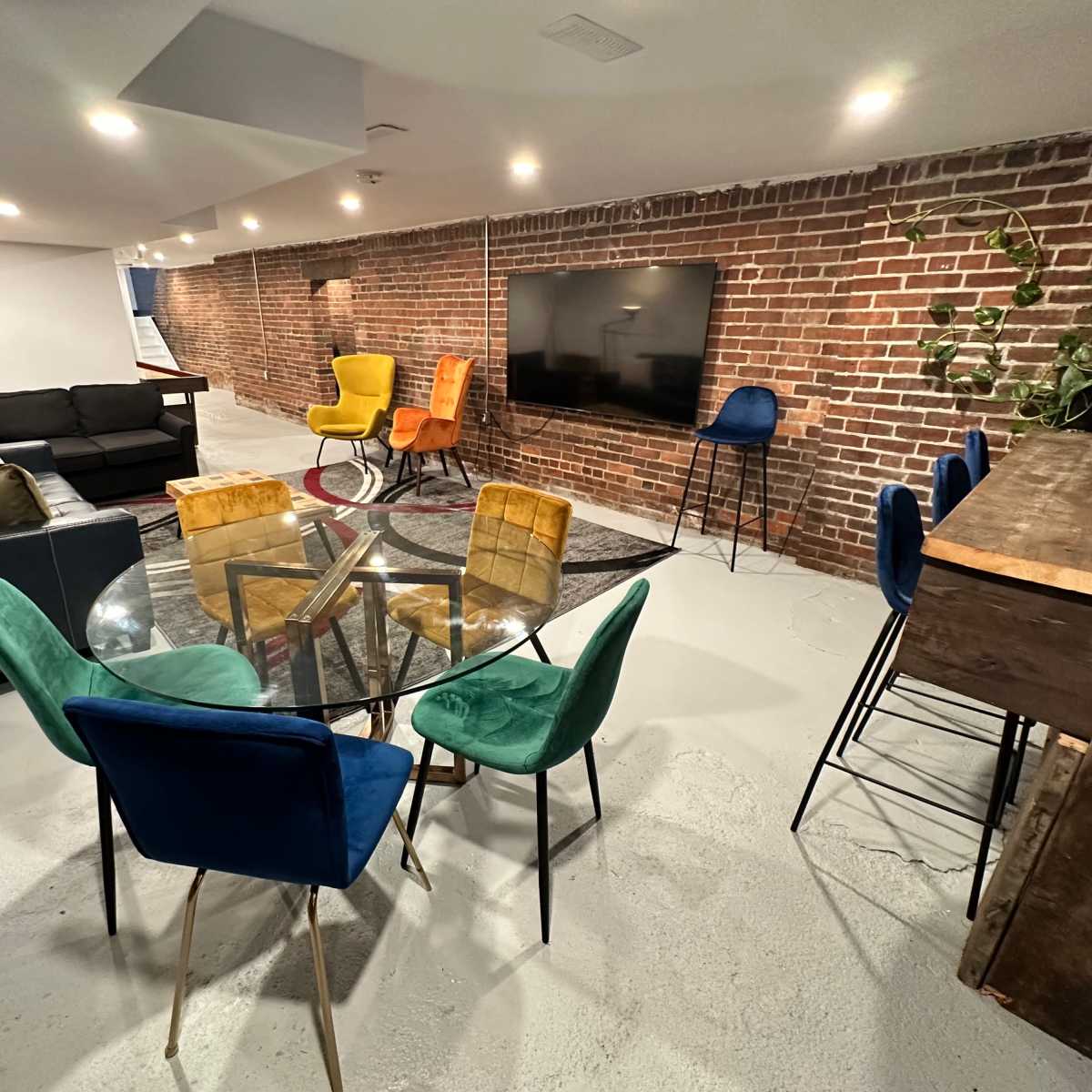 A modern basement living area with a seating arrangement that includes a black sofa, colorful chairs, a glass dining table, and a wall-mounted television.