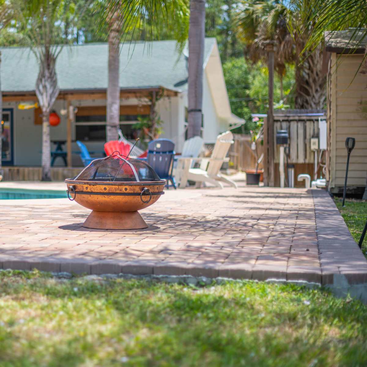 A decorative fire pit sits on a paved patio surrounded by palm trees and a swimming pool, with a house visible in the background.