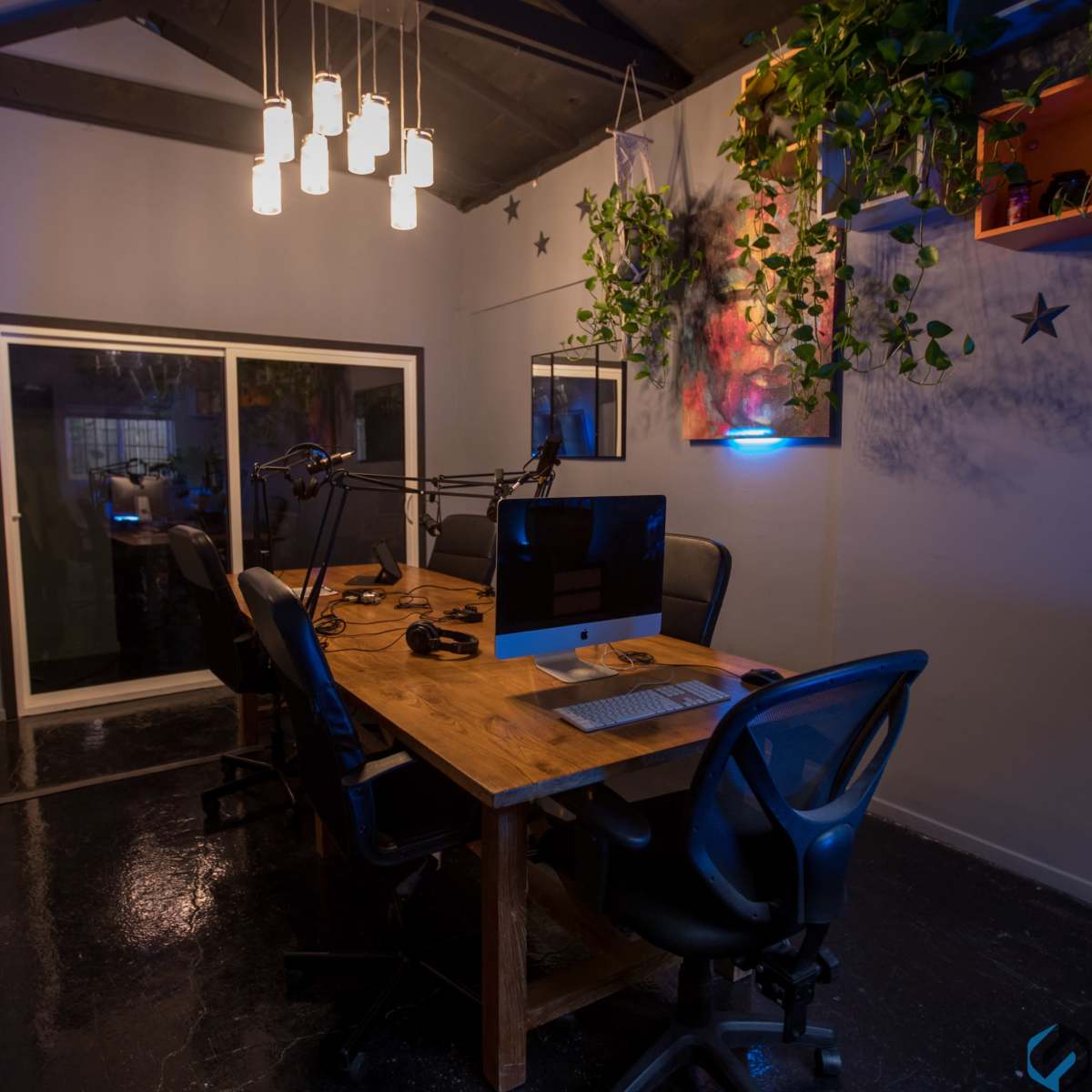 A modern office space features a wooden desk surrounded by black chairs, with hanging lights and plants adorning the walls.