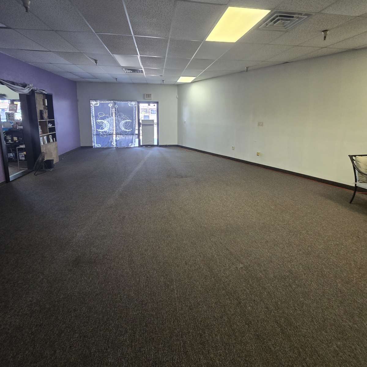 An empty commercial space with gray carpet and a purple accent wall, featuring a glass door leading outside.