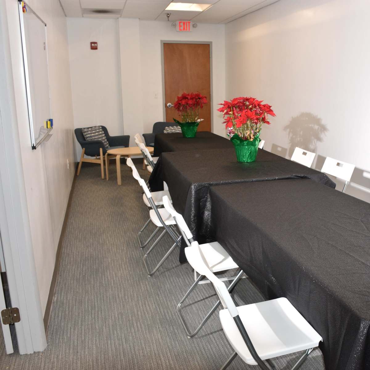 A long black-covered table with white chairs is arranged in a narrow meeting room, decorated with potted poinsettias at each end.