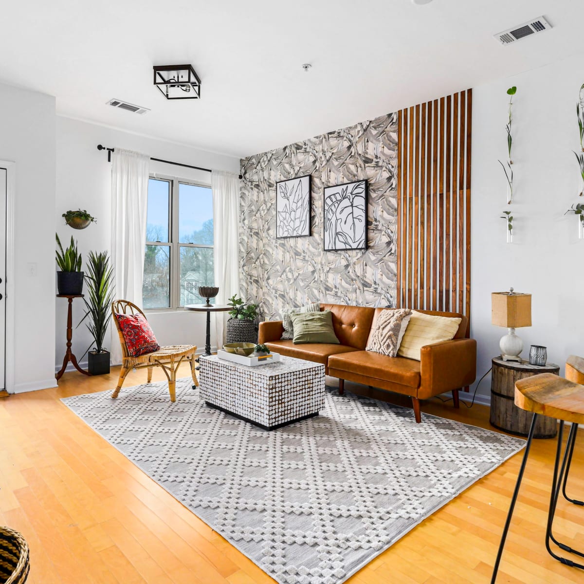 A modern living room features a brown leather couch, a decorative accent wall, and a window overlooking a balcony.