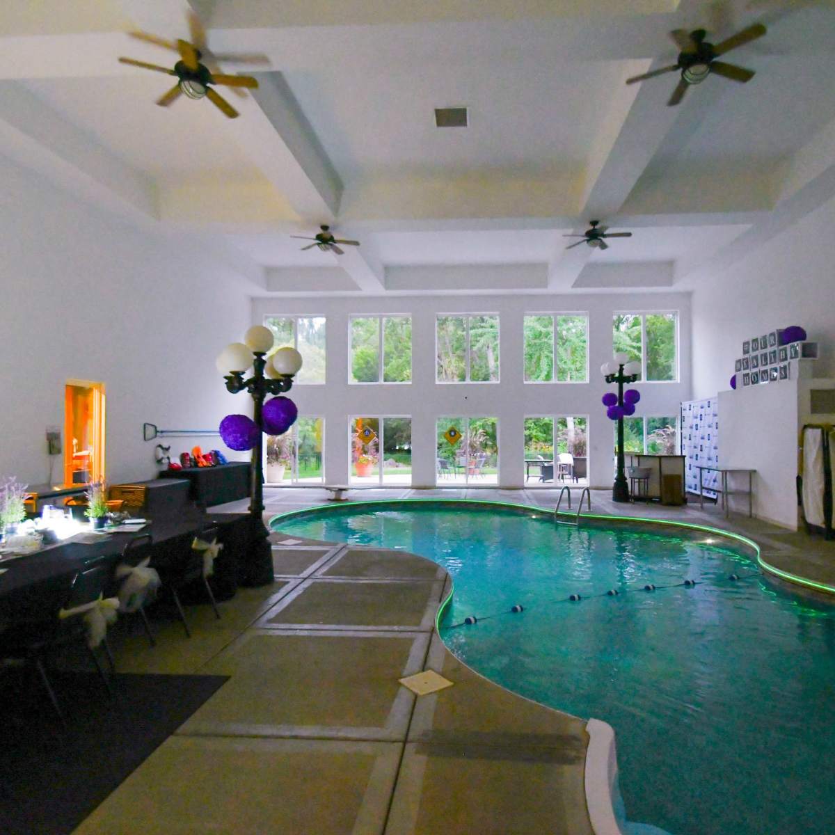 An indoor pool area with large windows, a dark table setup, and decorations in shades of purple.