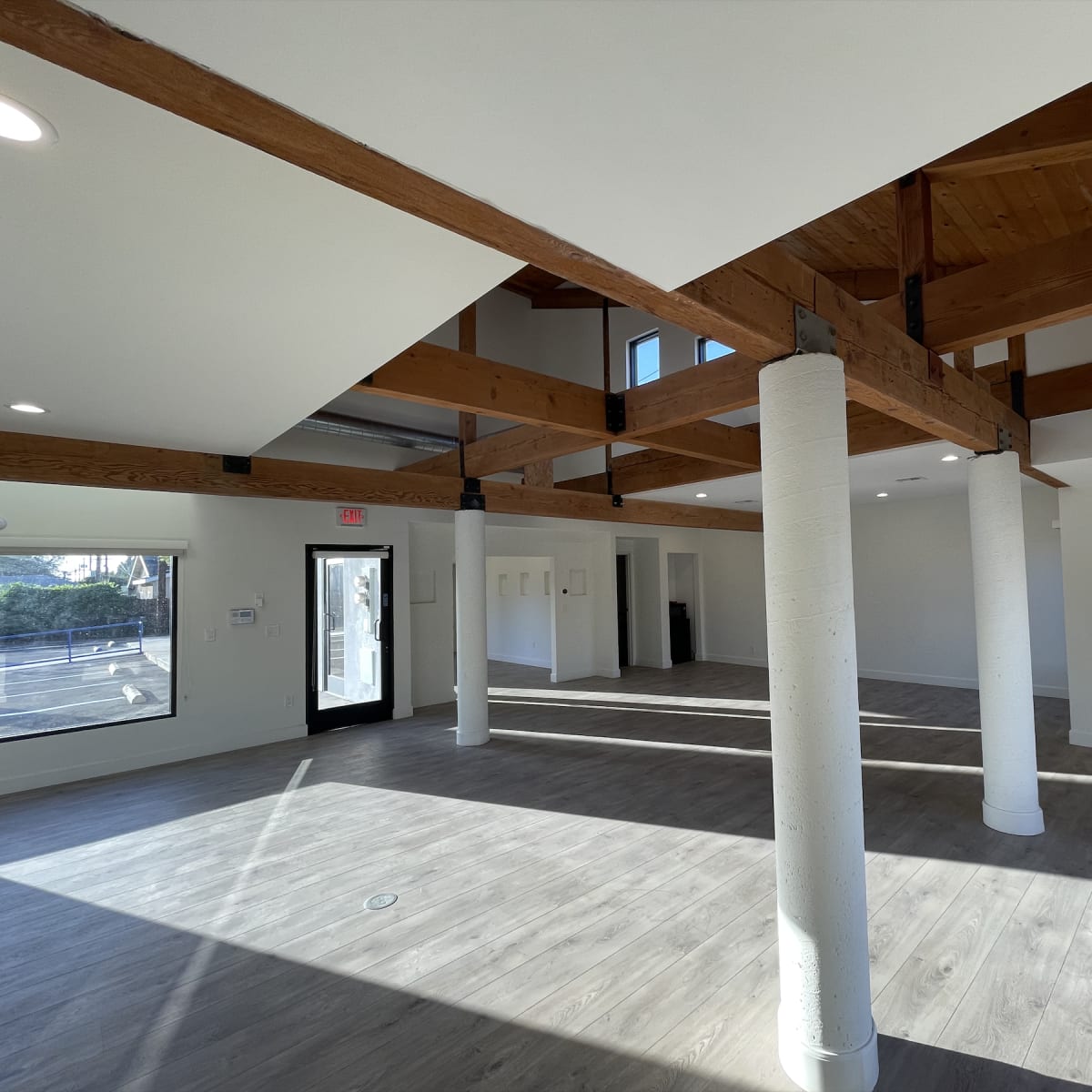 A spacious interior room with wooden beams, tall ceilings, and large windows, featuring a polished floor and columns.