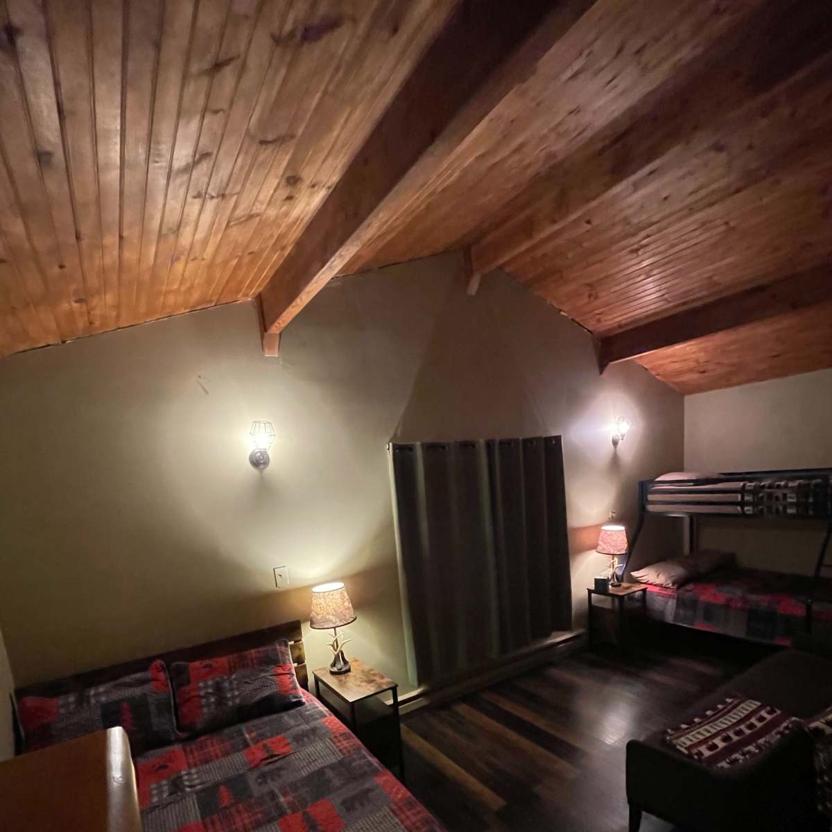 A dimly lit bedroom with two beds and a bunk bed, featuring wooden beams on the ceiling and a large window covered by curtains.