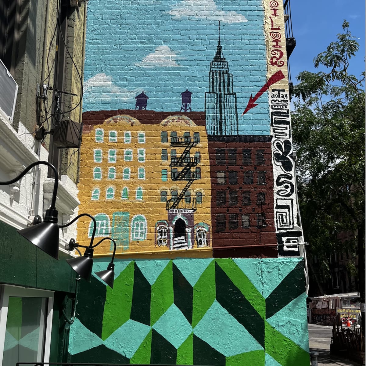 A colorful mural on a brick wall featuring buildings, an arrow pointing to the Empire State Building, and geometric green patterns below.