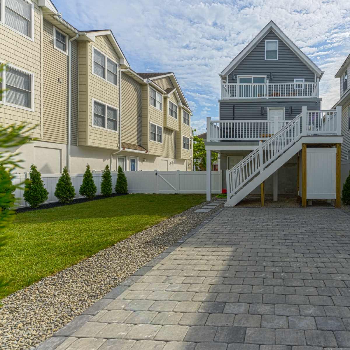 A two-story gray house with a front porch is situated in a paved backyard surrounded by decorative shrubs and adjacent homes.