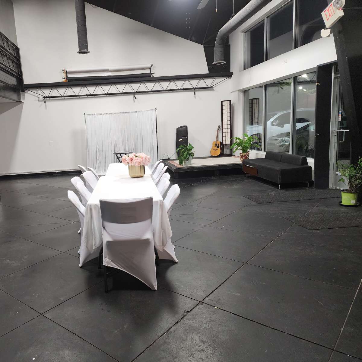 A spacious room with a long, white table set for dining, surrounded by chairs, and featuring a small stage or platform with a guitar in the corner.