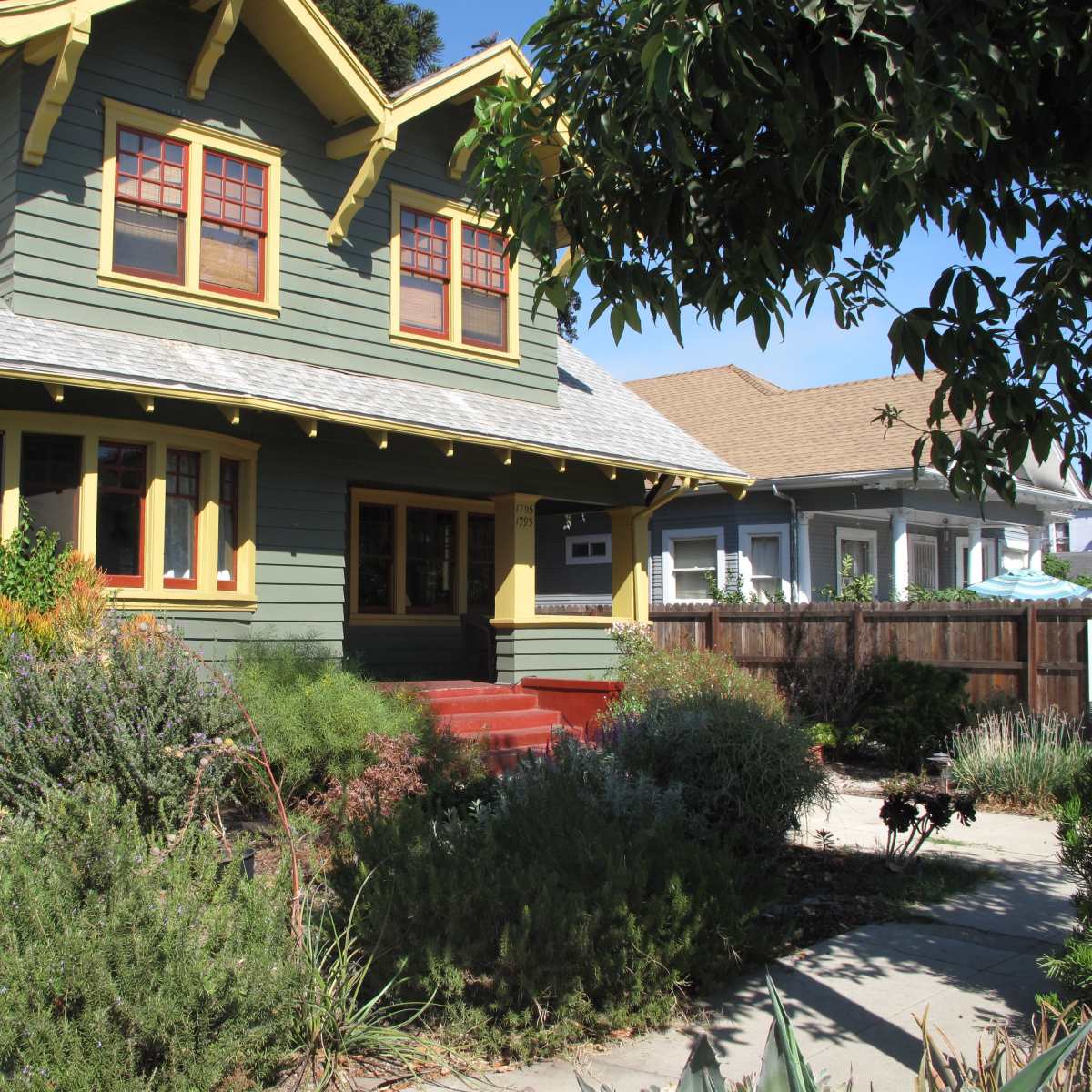 A two-story green house with yellow trim and a front porch is surrounded by various plants and shrubs.