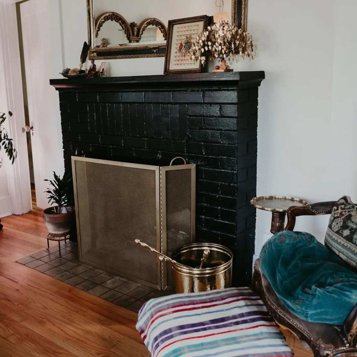 A black-painted fireplace with a decorative mirror above it, flanked by plants, a patterned stool, and an antique chair.
