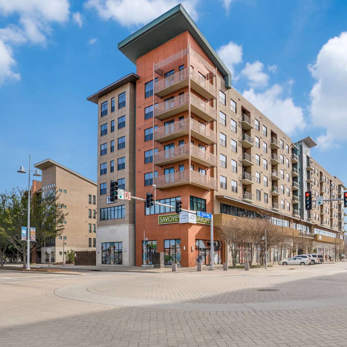 A modern multi-story building housing retail spaces at the ground level, located at a street intersection with traffic signals against a partly cloudy sky.