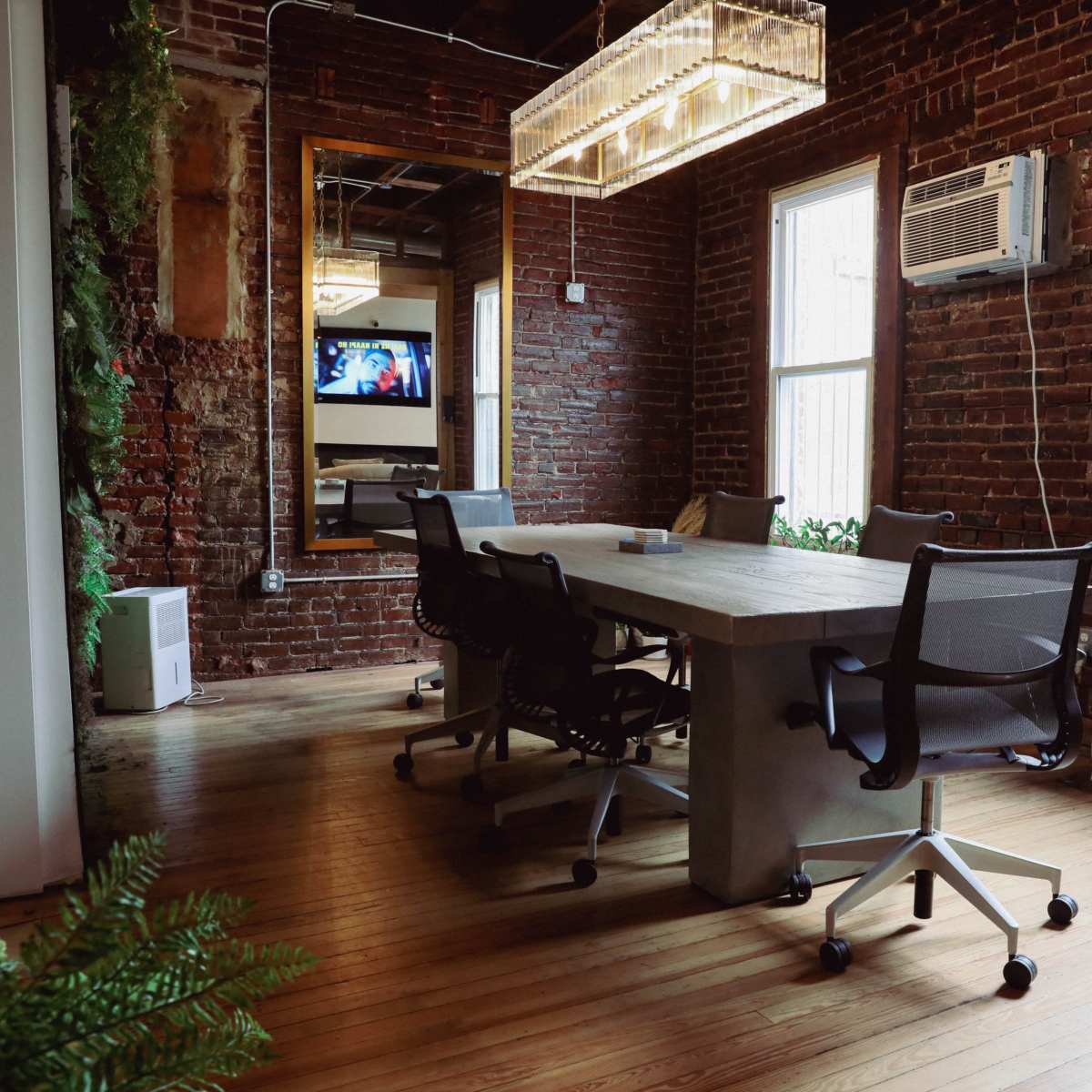 A conference room with a wooden table and several black office chairs, featuring exposed brick walls and large windows.