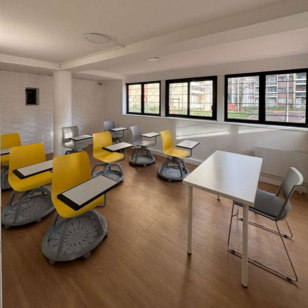 A bright classroom with several yellow and gray chairs arranged around white tables and large windows.