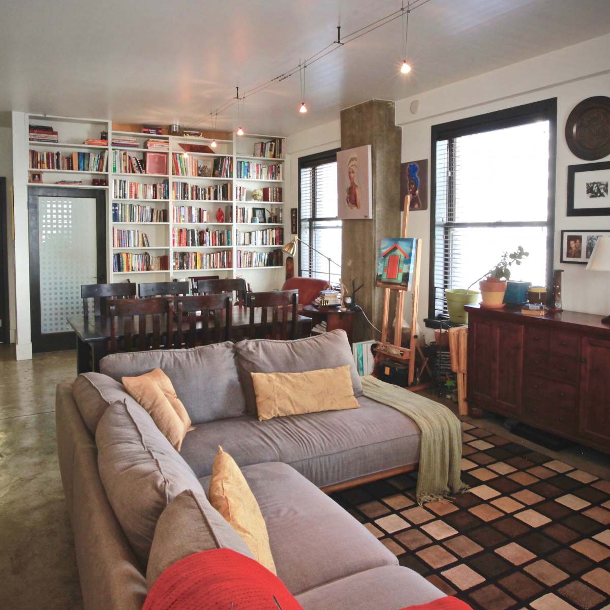 A modern living room with a gray sectional sofa, a large bookshelf filled with books, and an adjacent dining area with teal bar stools and a wooden dining table.