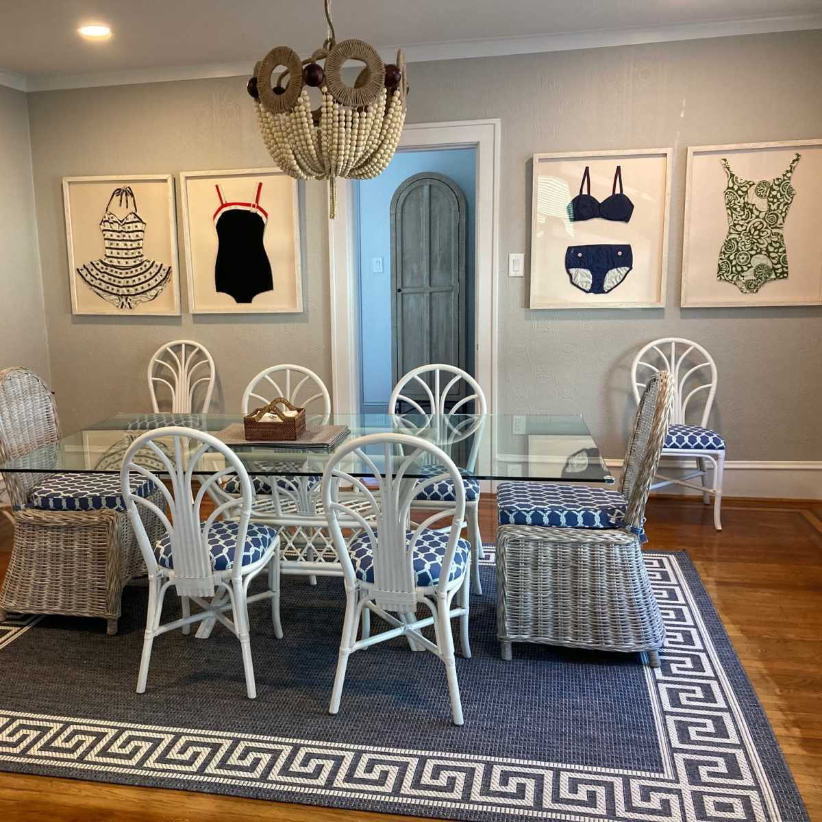 A dining room features a glass table surrounded by six white wicker chairs and two additional wicker chairs, with framed swimsuit artworks on the walls and a patterned rug underneath.